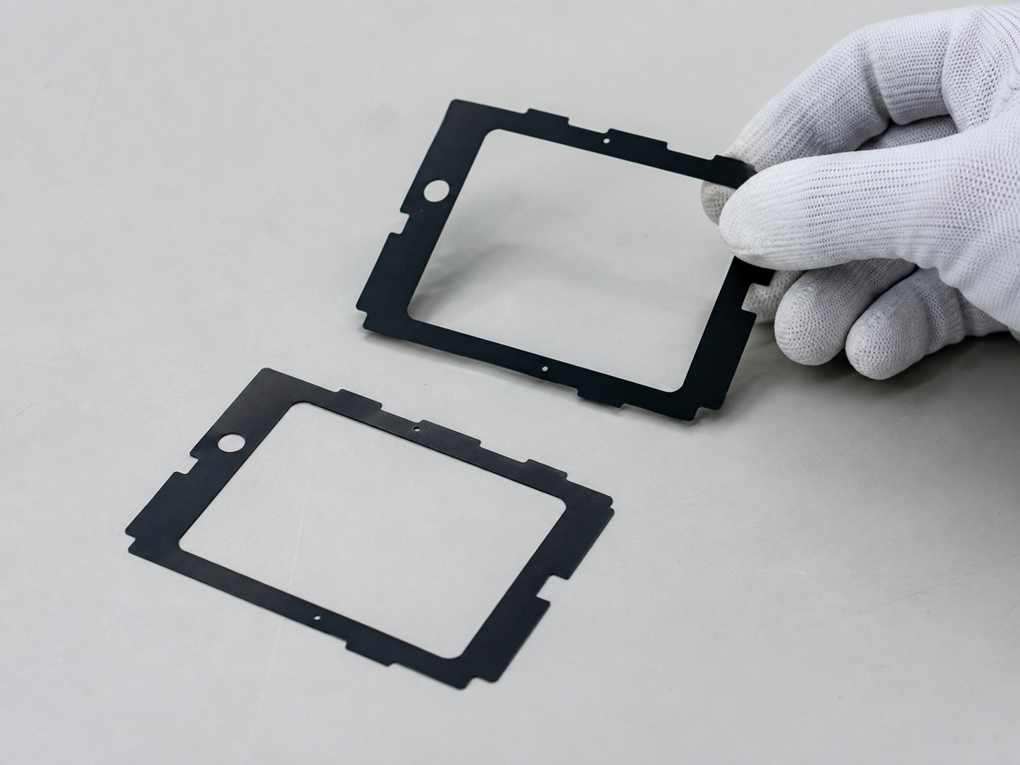 Image Description: Realistic factory quality inspection scene showing finished black PET light-blocking film components, adhesive-backed optical film frames, release liner sheets, inspection trays, digital calipers, thickness gauges, magnification tools, and organized OEM production batches. No complete electronic devices, displays, text, labels, logos, arrows, or icons. Image width: 1600–1920 px. Target file size: 100–200 KB.