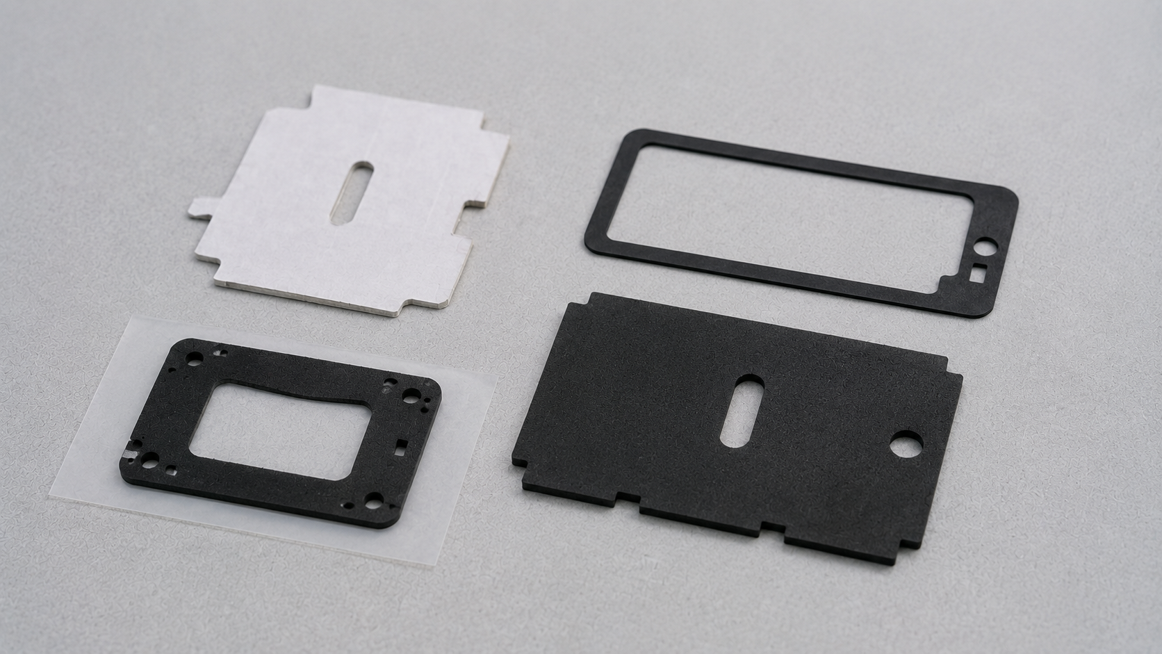 A realistic comparison between one simple hand-cut white paper-faced foam board prototype and several professionally die-cut black industrial foam components, including a narrow gasket frame and an adhesive-backed foam pad retained on a release liner, naturally positioned with slight irregular spacing on a clean light gray engineering work surface, realistic material textures, soft diffused lighting, generous negative space, no people, text, labels, logos, arrows, rulers, calipers, excessive tools or staged product display, 1600–1920 px wide, 100–200 KB