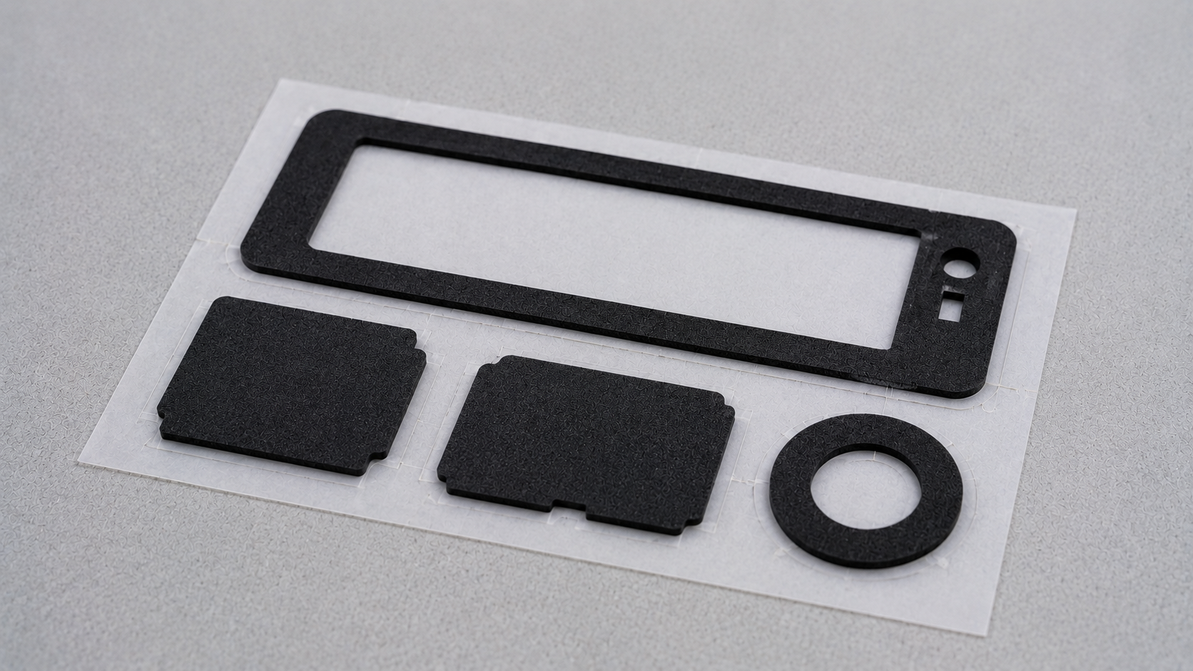 Several professionally die-cut black industrial foam components retained on a white release liner, including one narrow rectangular gasket frame, two small foam pads and one simple ring-shaped part, naturally arranged with realistic spacing and slight variation after clean waste-matrix removal, close industrial product photography on a light gray work surface, visible foam texture and clean cut edges, soft diffused lighting, no people, text, labels, logos, arrows, rulers, trays, calipers or excessive parts, 1600–1920 px wide, 100–200 KB