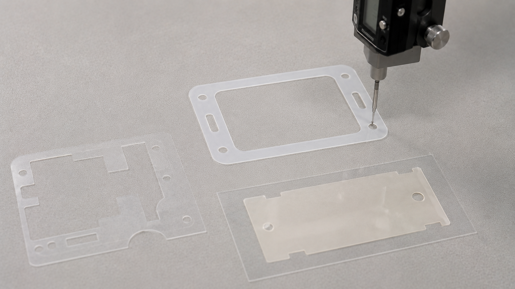 Realistic industrial quality inspection scene showing several custom die-cut PET electrical insulation film parts being checked on a clean light gray engineering work surface. Include one milky white PET insulation frame, one clear PET component with precision openings, and one adhesive-backed PET part retained on a release liner. Show a simple optical measuring instrument or digital caliper inspecting a critical opening or outside dimension. Keep the parts naturally spaced with realistic thin-film texture and clean die-cut edges. Professional OEM manufacturing environment, soft diffused lighting, subtle natural shadows, minimal composition, no stacked products, no excessive tools, no text, no labels, no logos, no arrows, no watermark, landscape 16:9.